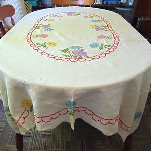 May include: White oval tablecloth with a red and white embroidered border and a floral design in the center. The flowers are pink, blue, yellow, and green.