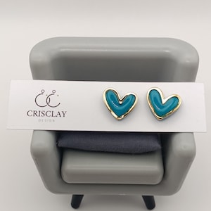 May include: A pair of heart-shaped stud earrings. The earrings feature a teal enamel heart design with a gold-colored border. The earrings are displayed on a white card with the text "CRISCLAY DESIGN".