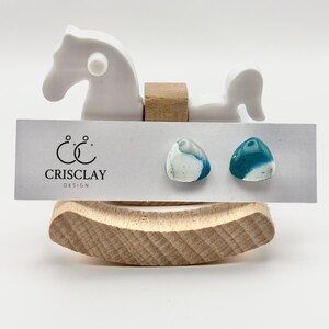 Può includere: Un paio di orecchini in ceramica color verde acqua e bianco con un design a spirale unico. Gli orecchini sono esposti su una carta bianca con il testo "Crisclay Design".