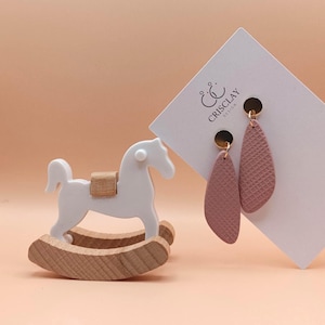Può includere: Un paio di orecchini pendenti rosa con texture e ganci dorati. Gli orecchini sono esposti su una carta bianca con il testo "CRISCLAY DESIGN" in nero.