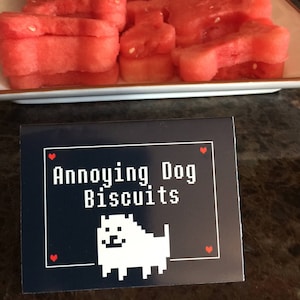 May include: A black and white sign with the text "Annoying Dog Biscuits" in white text. A pixelated white dog with black eyes is below the text. Two red hearts are on either side of the text.