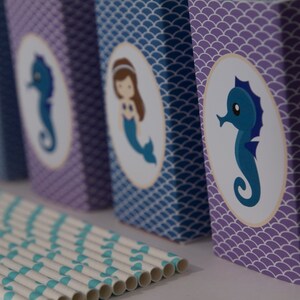 May include: Five purple and blue boxes with a mermaid or seahorse design. The boxes are decorated with a scale pattern. The boxes are lined up in a row. There are striped straws in front of the boxes.