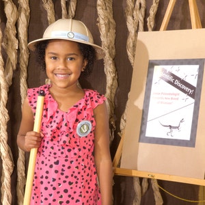 May include: A young person wearing a pink and black polka dot dress and a tan explorer hat. They are holding a wooden stick and looking at the camera. A sign behind them reads "Scientific Discovery! Junior Paleontologist Discovers New Breed of Dinosaur!"