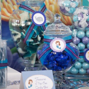 May include: A candy buffet table decorated with a mermaid theme. There are jars filled with blue and purple candies, and a sign that says "Treasures of the Sea Candy Bar".
