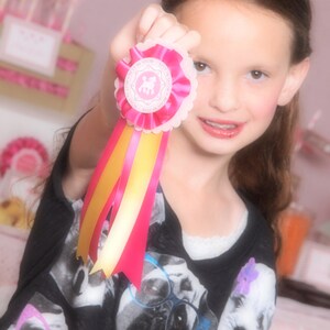 Peut inclure: Une jeune fille portant un t-shirt noir avec un imprimé de caniche blanc tient un ruban rose et jaune avec un cercle blanc qui dit "Best in Show" en lettres roses.