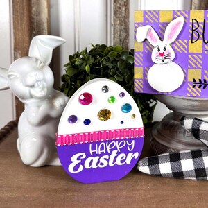 May include: Purple wooden Easter egg with a white background and colorful gems, a pink ribbon, and the text "Happy Easter". A purple wooden sign with a white bunny and yellow and white checkered background reads "Bunny Trail".