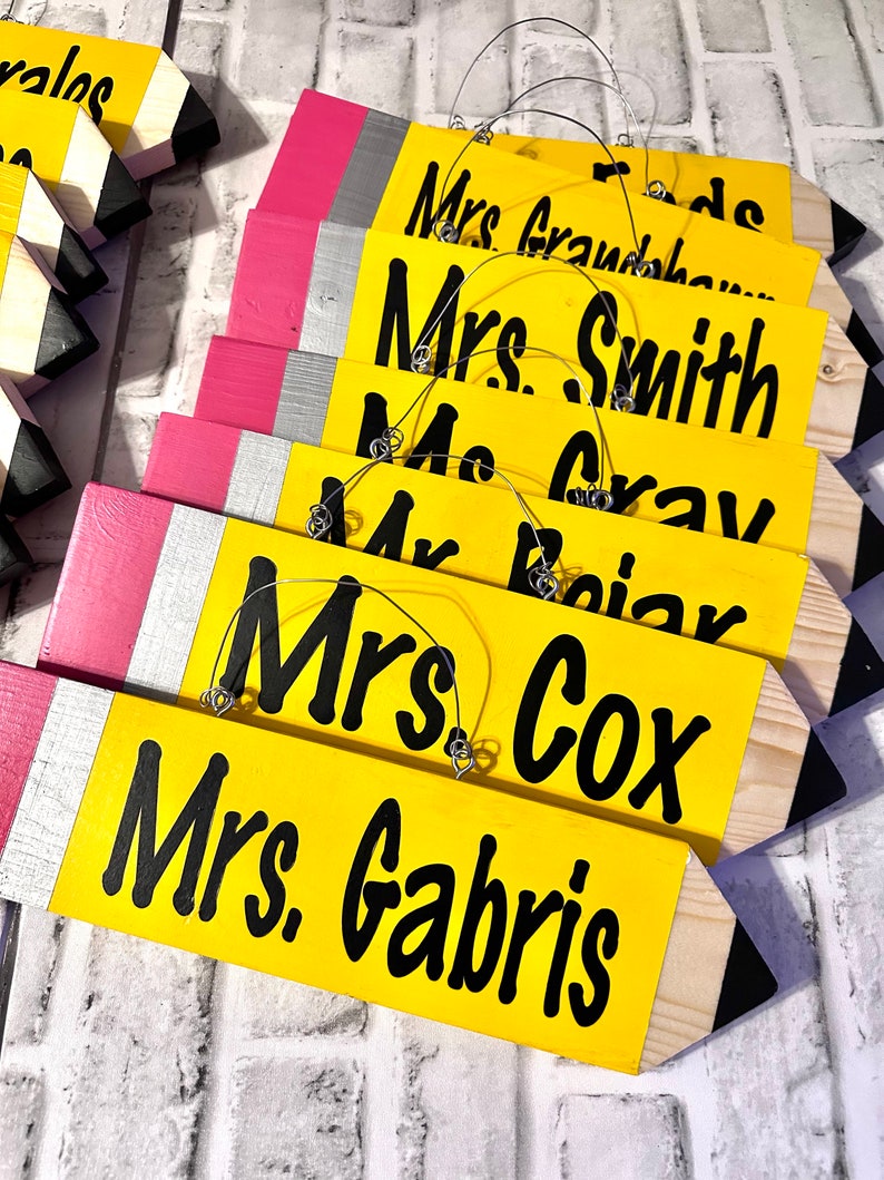 May include: A set of six yellow pencil-shaped wooden signs with black lettering. Each sign has a different name, including "Mrs. Gabris", "Mrs. Cox", "Mrs. Gray", "Mrs. Smith", and "Mrs. Grandhaw".