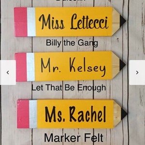 May include: Five wooden pencil-shaped signs with red and yellow paint. Each sign has a teacher's name written in black paint. The names are Ms. Schiding, Miss Lettecci, Mr. Kelsey, Ms. Rachel, and Mrs. Sexton. The signs are arranged vertically.
