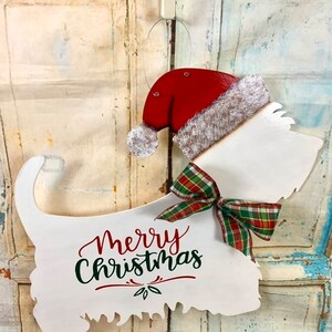 May include: White wooden cutout of a dog wearing a red Santa hat with a white fur trim and a green and red plaid bow. The dog is holding a sign that says "Merry Christmas" in green and red lettering.