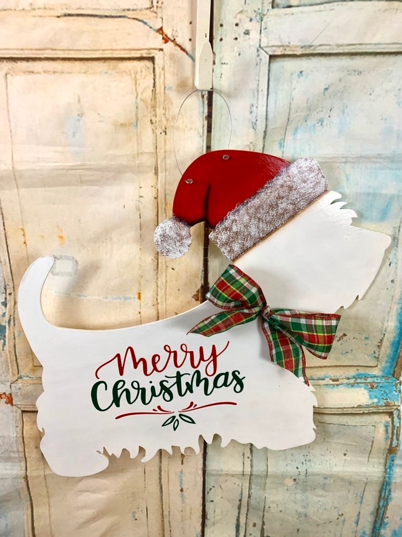 May include: White wooden cutout of a dog wearing a red Santa hat with a white fur trim and a green and red plaid bow. The dog is holding a sign that says "Merry Christmas" in green and red lettering.