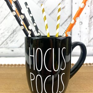 Puede incluir: Taza de cer&aacute;mica negra con la inscripci&oacute;n "Hocus Pocus" en blanco. La taza tiene un asa negra y est&aacute; llena de pajitas rayadas de color naranja, negro y dorado.