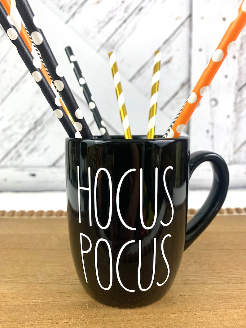 Puede incluir: Taza de cer&aacute;mica negra con la inscripci&oacute;n "Hocus Pocus" en blanco. La taza tiene un asa negra y est&aacute; llena de pajitas rayadas de color naranja, negro y dorado.