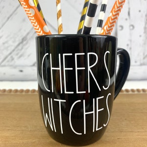 Puede incluir: Taza de cer&aacute;mica negra con una inscripci&oacute;n blanca que dice "Cheers Witches". La taza est&aacute; llena de pajitas rayadas de colores.