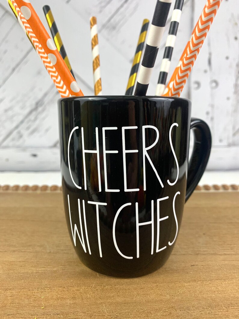Puede incluir: Taza de cer&aacute;mica negra con una inscripci&oacute;n blanca que dice "Cheers Witches". La taza est&aacute; llena de pajitas rayadas de colores.