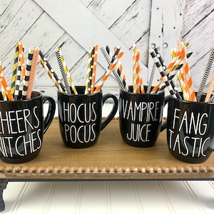 Puede incluir: Cuatro tazas negras con letras blancas en una bandeja de madera. Las tazas est&aacute;n llenas de pajitas rayadas de colores. Las tazas dicen "Cheers Witches", "Hocus Pocus", "Vampire Juice" y "Fangtastic".