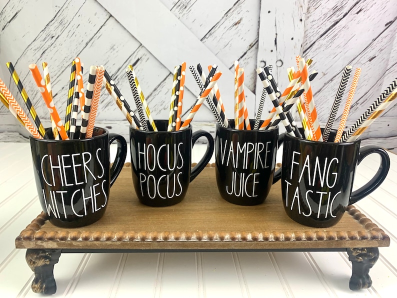 Puede incluir: Cuatro tazas negras con letras blancas en una bandeja de madera. Las tazas est&aacute;n llenas de pajitas rayadas de colores. Las tazas dicen "Cheers Witches", "Hocus Pocus", "Vampire Juice" y "Fangtastic".