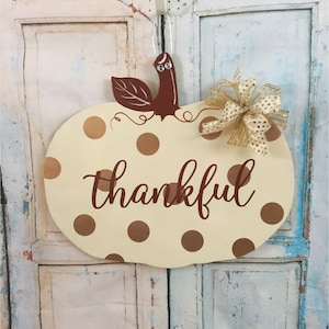 May include: A cream-colored pumpkin-shaped door hanger with brown polka dots and the word "thankful" written in brown. A brown stem with a leaf and a gold bow are attached to the top.