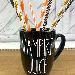 Puede incluir: Taza de cer&aacute;mica negra con asa blanca y el texto "Vampire Juice" en letras blancas. La taza est&aacute; llena de pajitas de papel a rayas naranja, negras y doradas.