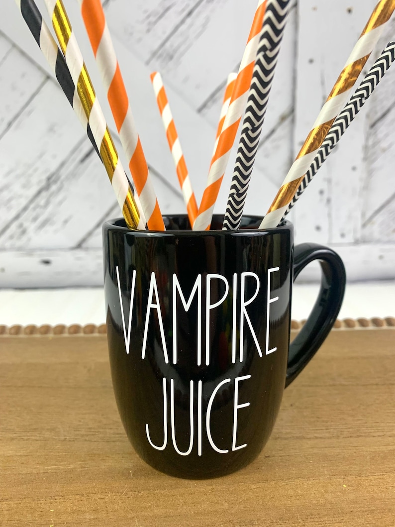 Puede incluir: Taza de cer&aacute;mica negra con asa blanca y el texto "Vampire Juice" en letras blancas. La taza est&aacute; llena de pajitas de papel a rayas naranja, negras y doradas.