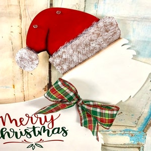 May include: White wooden Christmas ornament shaped like a dog wearing a red Santa hat with a white fur trim and a green and red plaid bow. The ornament says "Merry Christmas" in red and green lettering.
