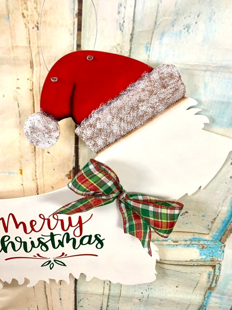 May include: White wooden Christmas ornament shaped like a dog wearing a red Santa hat with a white fur trim and a green and red plaid bow. The ornament says "Merry Christmas" in red and green lettering.