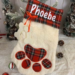 May include: Red and white plaid Christmas stocking with a white faux fur body, a paw print design, and the name "Phoebe" embroidered on the cuff.