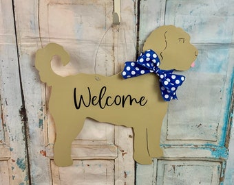 May include: A tan wooden dog silhouette with a blue and white polka dot bow tie. The dog is standing on a wooden door and says "Welcome".