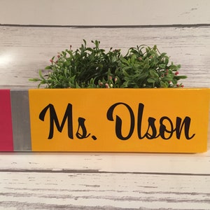 May include: A yellow pencil-shaped wooden sign with a red and silver band at the bottom. The sign says "Ms. Olson" in black cursive lettering.
