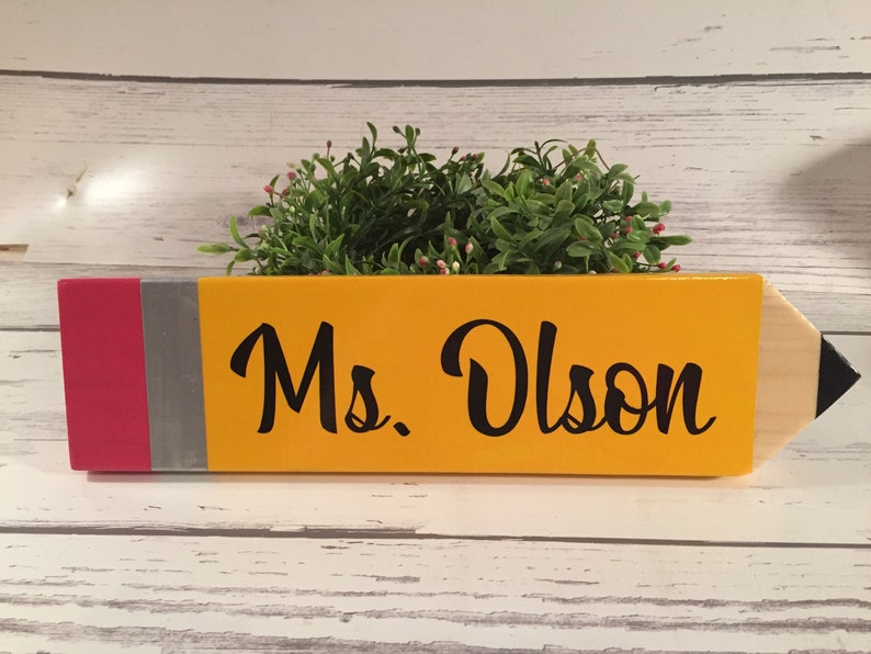 May include: A yellow pencil-shaped wooden sign with a red and silver band at the bottom. The sign says "Ms. Olson" in black cursive lettering.