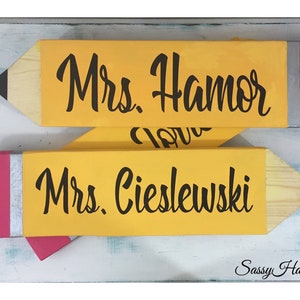 May include: Two wooden pencil-shaped signs with pink erasers. The top sign reads "Mrs. Hamor" and the bottom sign reads "Mrs. Cieslewski". The signs are on a light blue background with the text "Sassy Hang Ups" in the bottom right corner.