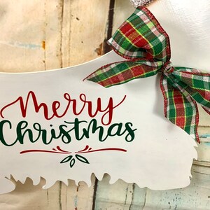 May include: White wooden sign shaped like a Christmas tree with the text "Merry Christmas" in red and green lettering. A red and green plaid bow is attached to the top of the sign.