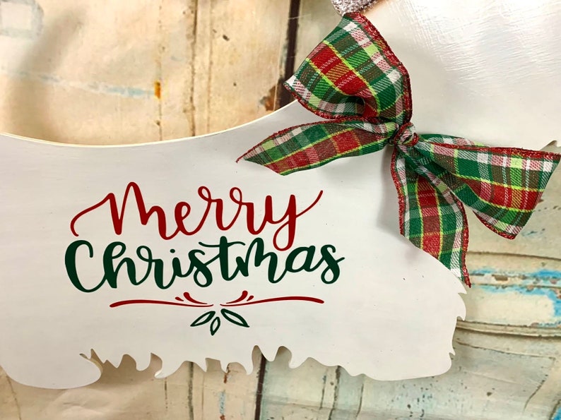 May include: White wooden sign shaped like a Christmas tree with the text "Merry Christmas" in red and green lettering. A red and green plaid bow is attached to the top of the sign.