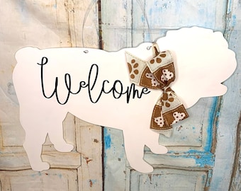 May include: White wooden door hanger shaped like a dog with the word "Welcome" written in black script. A brown and white bow with paw print design is tied around the dog's neck.