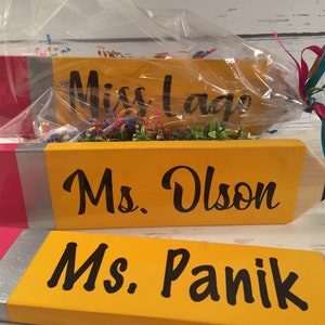 May include: Three yellow pencil-shaped wooden signs with pink and silver accents. The signs are personalised with the names "Miss Lage", "Ms. Olson", and "Ms. Panik".
