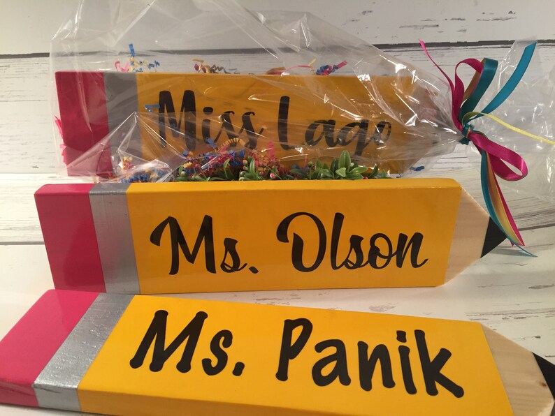 May include: Three yellow pencil-shaped wooden signs with pink and silver accents. The signs are personalised with the names "Miss Lage", "Ms. Olson", and "Ms. Panik".