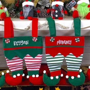 May include: Two green and red elf stocking holders with white stripes, personalized with the names "Mommy" and "Reygan". The stockings are hanging on a white mantel with a Christmas tree in the background.