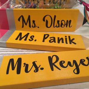May include: Three yellow and pink wooden pencil-shaped signs with black lettering. The signs read "Ms. Olson", "Ms. Panik", and "Mrs. Reyes".