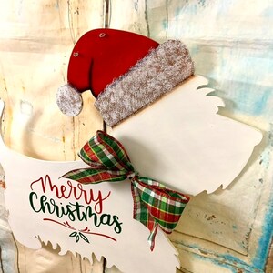 May include: White wooden Christmas door hanger shaped like a dog wearing a red Santa hat with a green and red plaid bow. The dog is wearing a Santa hat and the words "Merry Christmas" are written on the dog's body.