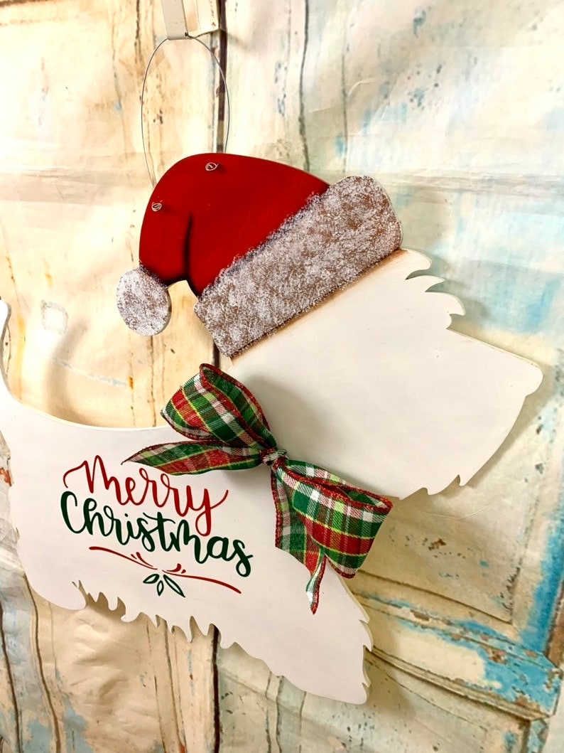 May include: White wooden Christmas door hanger shaped like a dog wearing a red Santa hat with a green and red plaid bow. The dog is wearing a Santa hat and the words "Merry Christmas" are written on the dog's body.