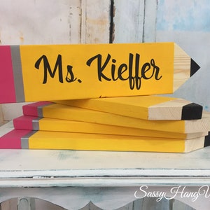 May include: A stack of wooden pencil-shaped signs with a pink and grey eraser detail. The top sign is painted yellow and has the text "Ms. Kieffer" in black.