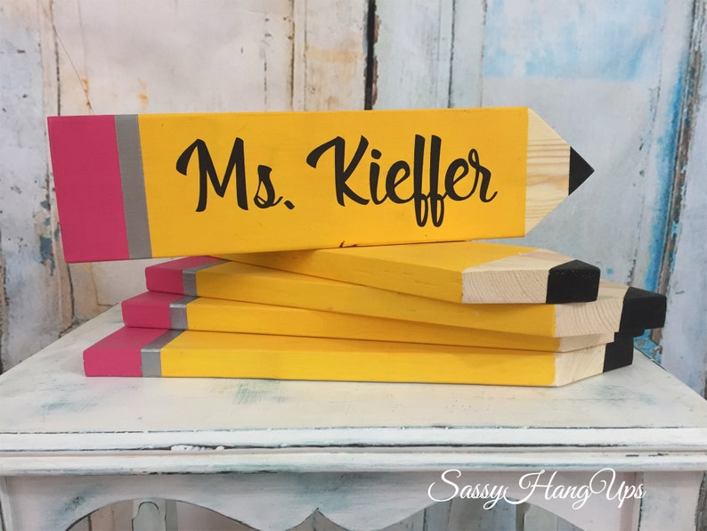 May include: A stack of wooden pencil-shaped signs with a pink and grey eraser detail. The top sign is painted yellow and has the text "Ms. Kieffer" in black.