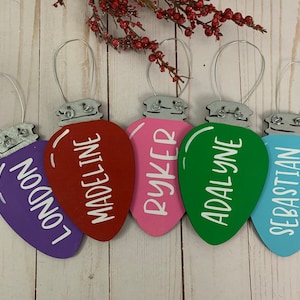May include: Five wooden lightbulb ornaments with silver wire hangers. Each ornament is painted a different color and has a name written on it in white paint: London, Madeline, Ryker, Adalyn, and Sebastian.