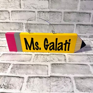 May include: A yellow wooden pencil-shaped sign with a pink and grey stripe at the top. The sign has a metal hanger and says "Ms. Galati" in black lettering.
