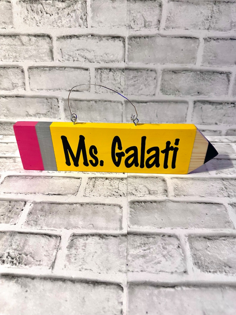 May include: A yellow wooden pencil-shaped sign with a pink and grey stripe at the top. The sign has a metal hanger and says "Ms. Galati" in black lettering.