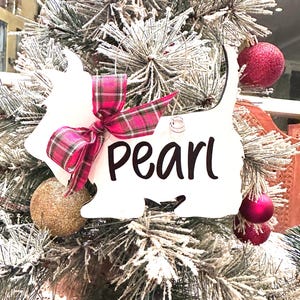 May include: A white Scottie dog Christmas ornament with the name "Pearl" in black script. The ornament is adorned with a pink and red plaid bow and hangs on a frosted Christmas tree with red and gold ornaments.