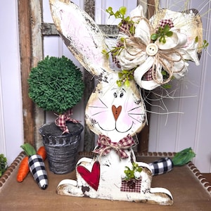 May include: A white wooden bunny rabbit with a red heart, a red and white gingham bow, and a white bow with a burlap bow on top. The bunny is sitting on a wooden surface with two black and white checkered carrots.
