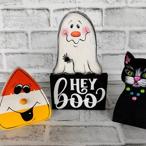 May include: Halloween decorations featuring a ghost, a black cat, and a candy corn. The ghost is white with a black spider on its chest. The cat is black with white accents and pink ears. The candy corn is yellow, orange, and white with a smiling face. The decorations are all painted on wood and have a rustic look.