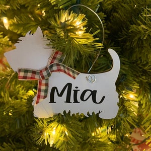 May include: White wooden Christmas ornament shaped like a dog with a red and white plaid bow and the name "Mia" written in black.