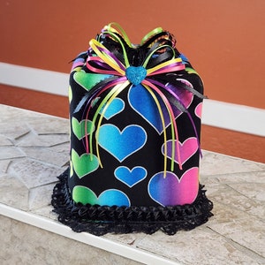 May include: A black tissue box cover with a rainbow heart pattern and a black lace trim. The cover has a large bow with multi-colored ribbons.