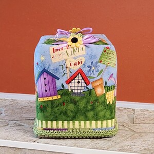 May include: A decorative fabric box with a blue sky background, featuring birdhouses, flowers, and the text "Friends Always Welcome". The box is trimmed with green and yellow accents and a sunflower bow.